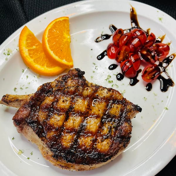 Grilled pork chop with orange slices and cherry tomatoes drizzled with balsamic glaze on a white plate.