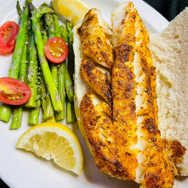 Seasoned fish fillet on a bun with asparagus, cherry tomatoes, and lemon wedges on a white plate.